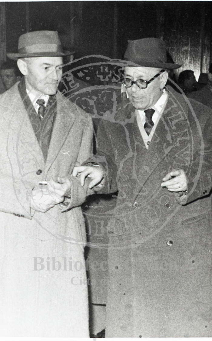 Primo piano di due uomini in abiti civili: l'uomo a destra è il politico Umberto Terracini. Indossano due cappotti e l'uomo a sinistra tiene in mano una sigaretta. Alle loro spalle altre persone