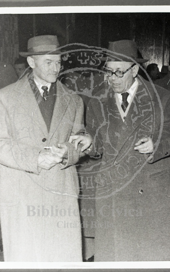 Primo piano di due uomini in abiti civili: l'uomo a destra è il politico Umberto Terracini. Indossano due cappotti e l'uomo a sinistra tiene in mano una sigaretta. Alle loro spalle altre persone